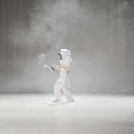 Une personne en tenue d'apicultueur est dans un environnement grisonnant, il a un objet faisant de la fumée à la main